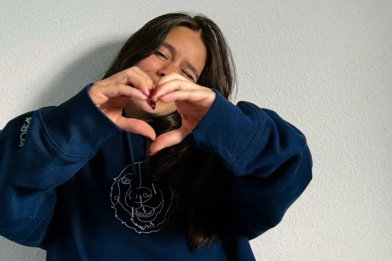 Model wearing a Lnnya blue hoodie with a bear graphic, making a heart shape with their hands against a plain background.