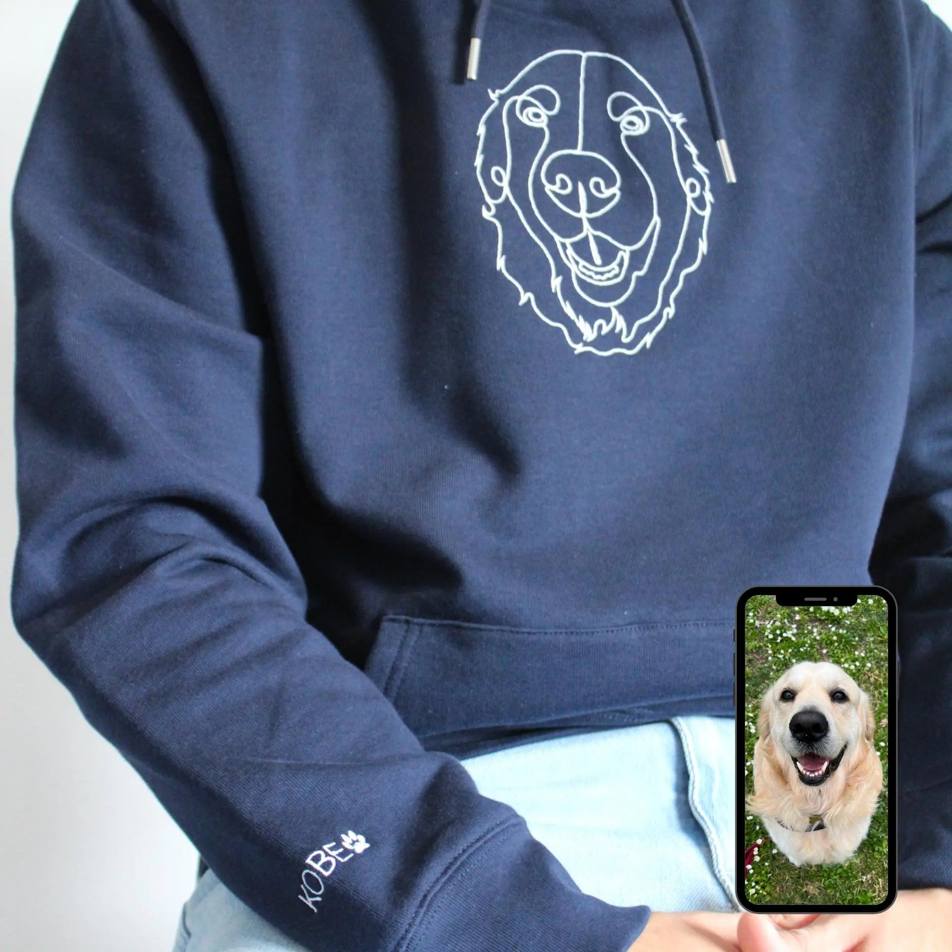 Navy blue hoodie with a white dog design on the chest, held by a person, with a phone showing a dog photo.