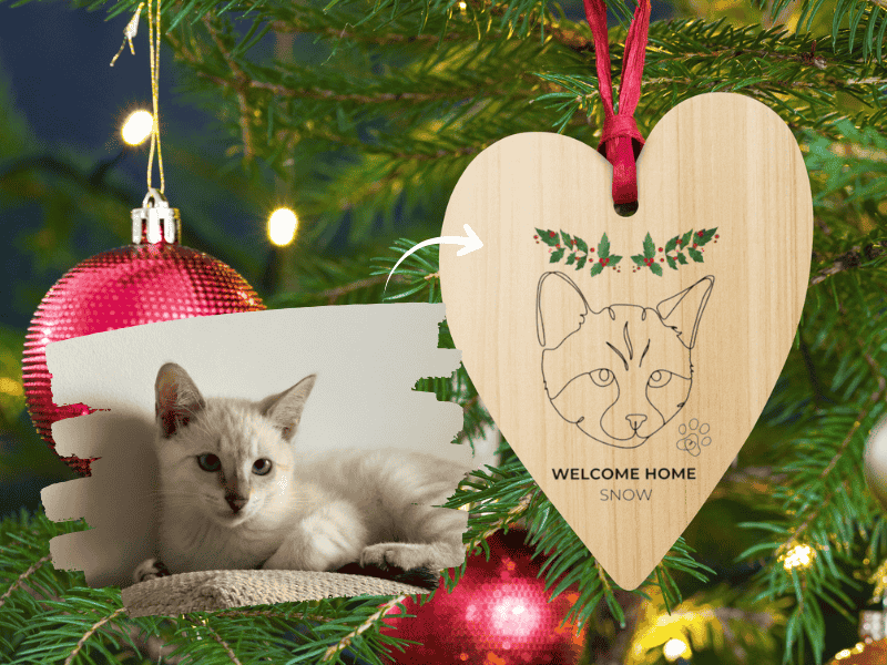 Heart-shaped wooden ornament with a cat illustration and 'Welcome Home Snow' text, hanging on a Christmas tree.