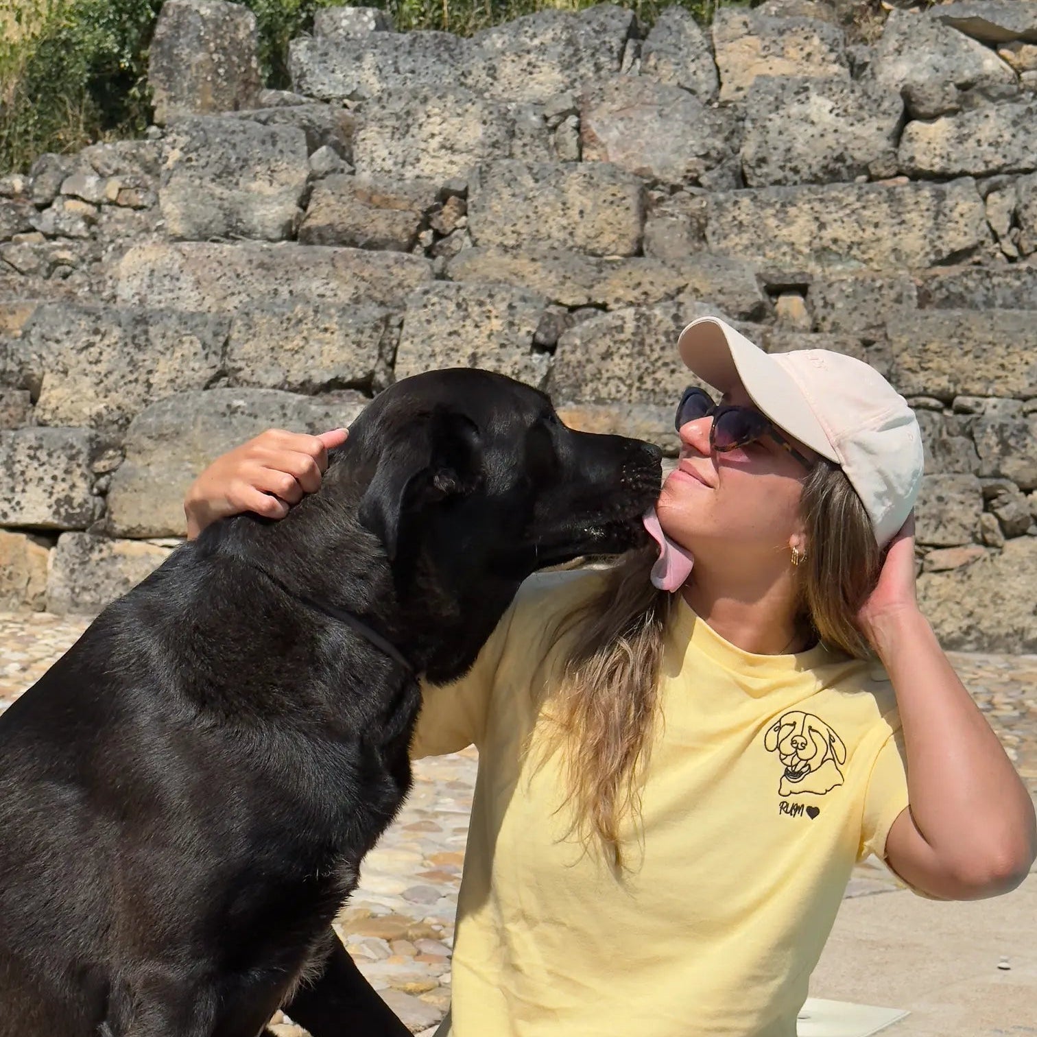 Woman in yellow shirt and cap sitting with a black dog in front of stone ruins
