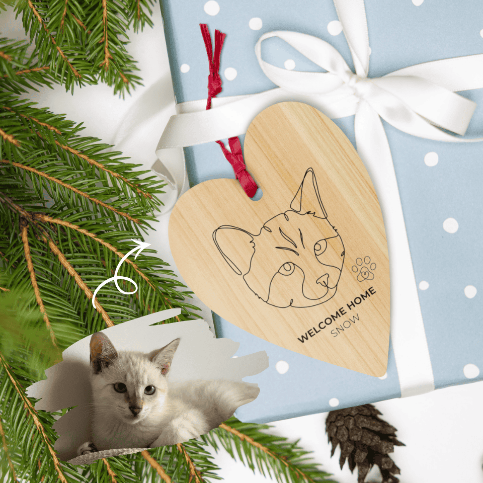 Wooden heart-shaped ornament with a cat design and 'Welcome Home Snowy' text, surrounded by Christmas decorations.