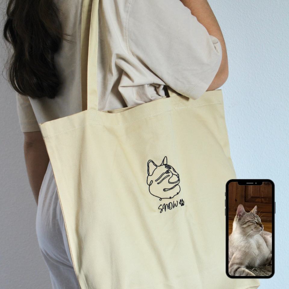 Person holding a beige tote bag with a cat illustration and 'Snow' text, next to a phone showing a cat photo.