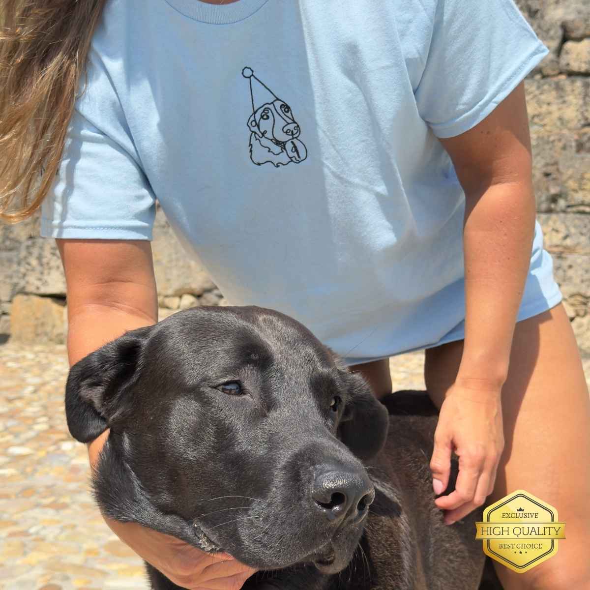 Person wearing a light blue t-shirt with a dog graphic, standing next to a black dog outdoors.