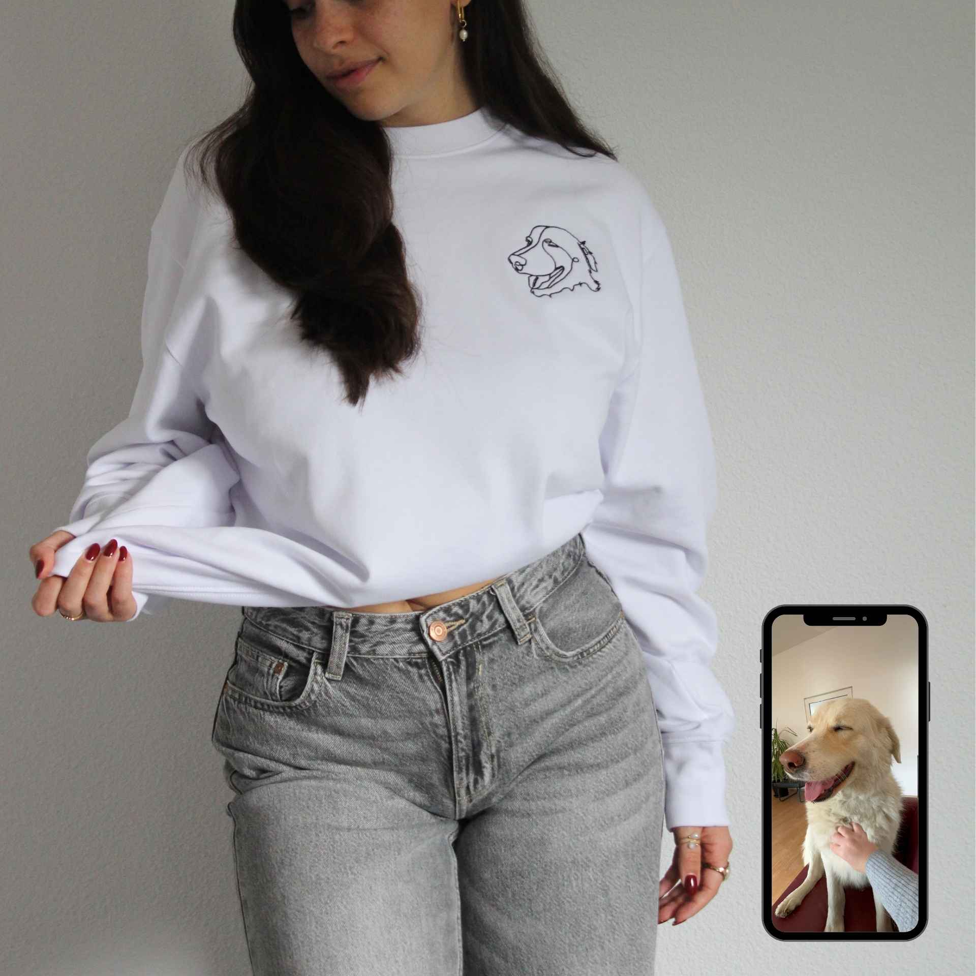 Person wearing a white sweatshirt with a dog design and gray jeans, holding a phone with a dog photo.