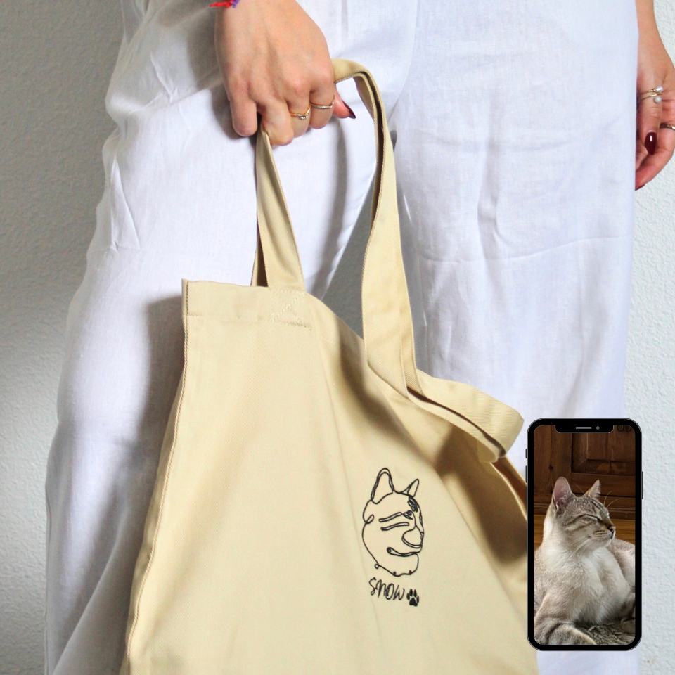 Beige tote bag with a cat illustration held by a person wearing a white outfit.