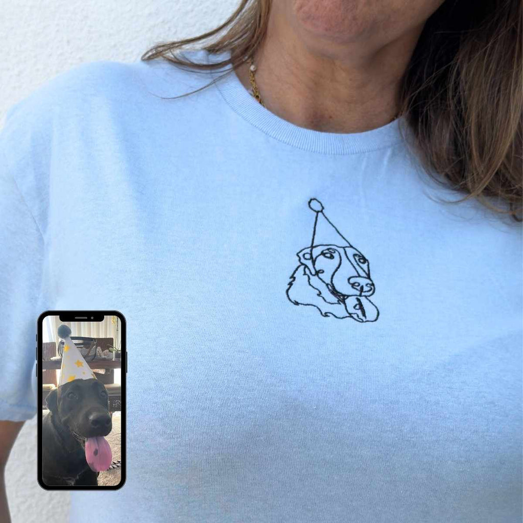 Person wearing a light blue t-shirt with a dog graphic and a phone displaying a photo of a dog wearing a party hat.
