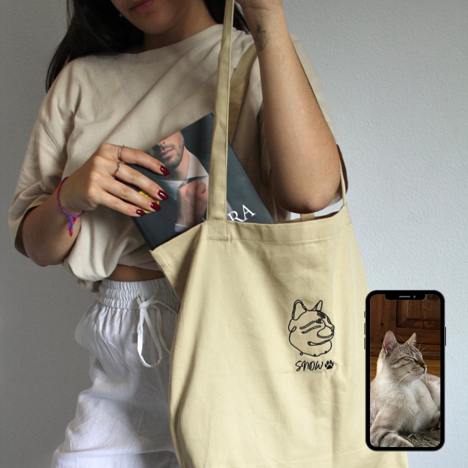 Person holding a beige tote bag with a cat design, wearing a white shirt and white pants.