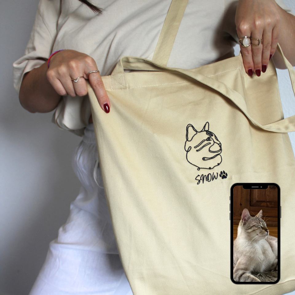 Beige tote bag with a cat illustration and text, held by a person.