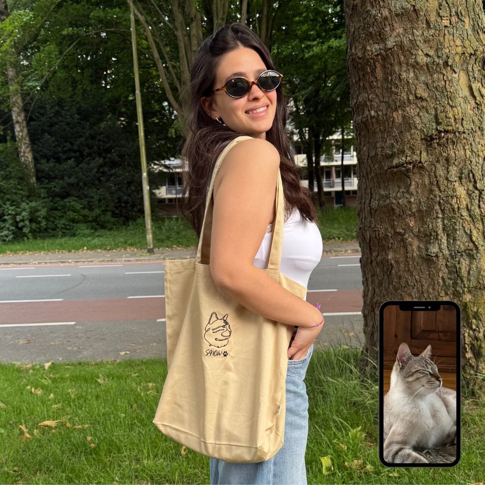 Woman holding a beige tote bag with a cat design, standing outdoors near a tree.
