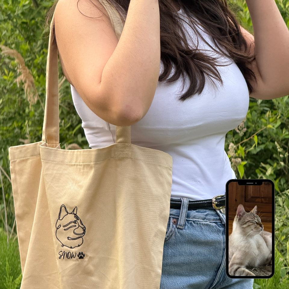 Person holding a beige tote bag with a cat design, standing outdoors.