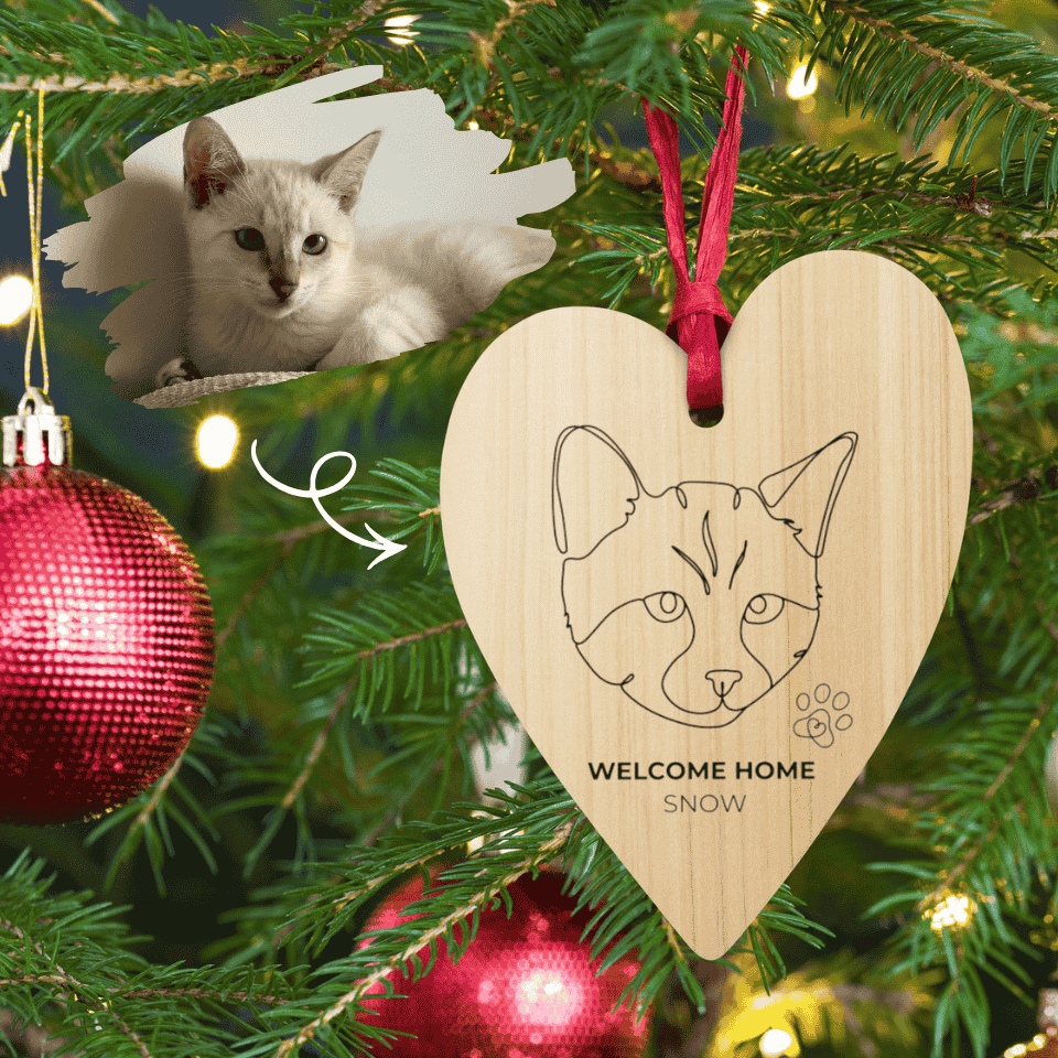 Wooden heart-shaped ornament with a cat's face and 'Welcome Home Snow' text, hanging on a Christmas tree.