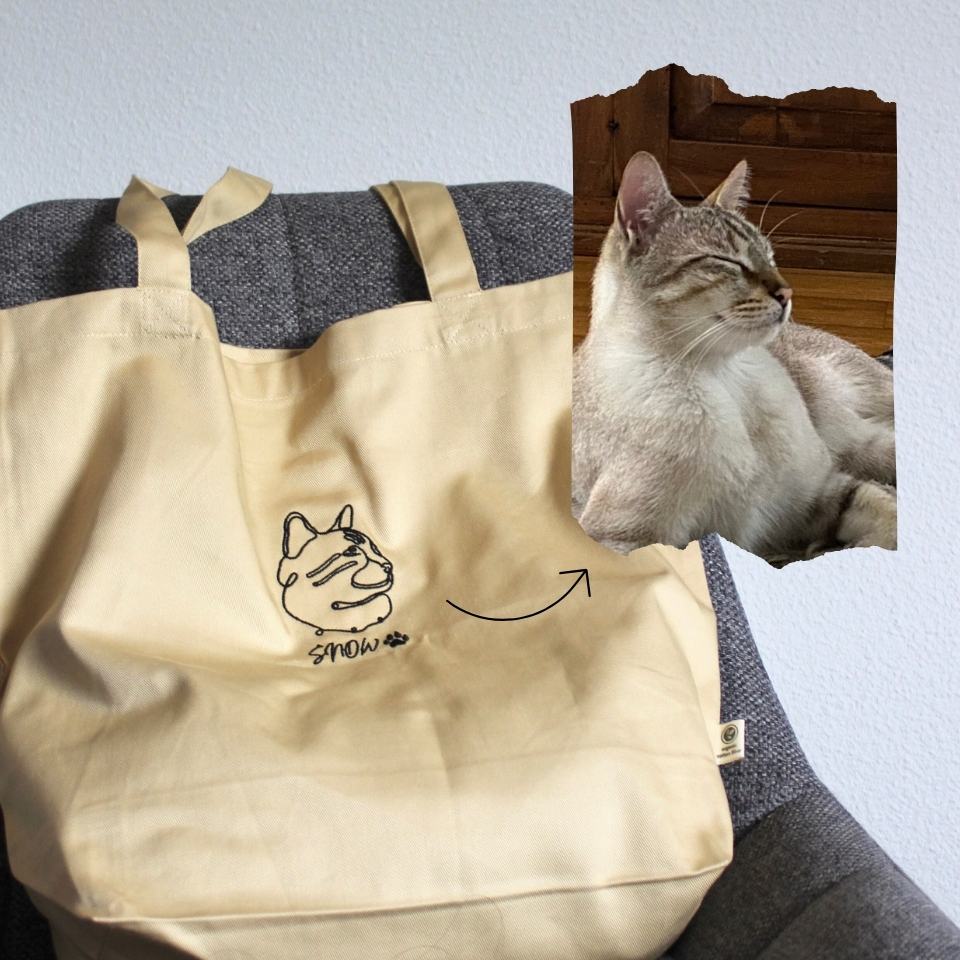 Beige tote bag with a cat illustration and text, placed on a surface with a cat peeking from behind.