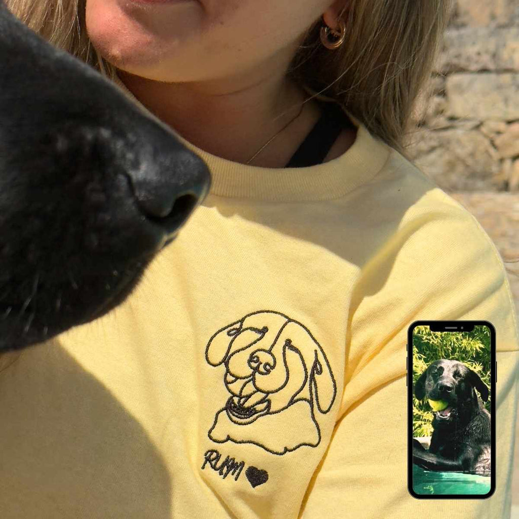 Person wearing a yellow sweatshirt with a dog graphic, holding a phone showing a photo of a dog.